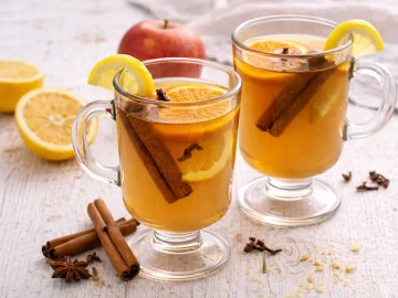 Apfelpunsch in zwei Glasbechern mit Zimtstange, Nelken und Zitronenscheibe auf weißem Holzhintergrund