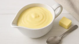 Helle Eiersoße in einer weißen Sauciere auf weißem Holzhintergrund, daneben ein Löffel und etwas Butter.