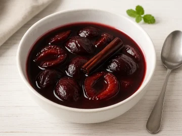 Weiße Schüssel mit feinem Pflaumenkompott, verfeinert mit Rotwein und Zimtstange auf weißem Holzhintergrund