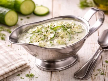 Gurken-Sahne-Soße mit Gurkenscheiben in Edelstahl-Sauciere auf weißem Holztisch angerichtet