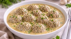 Heringsklopse in cremiger Senfsoße in einer weißen Servierschale mit hohem Rand auf hellem Holztisch