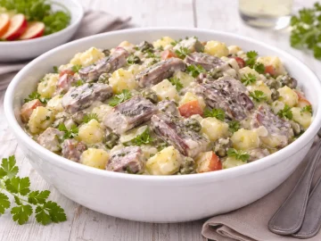 Weißer Teller mit Heringssalat aus Heringsstreifen, Kartoffelwürfeln, Apfel und Gurke auf weißem Holzhintergrund mit sichtbarer Maserung