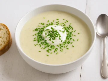 Kalte Kartoffelkremsuppe mit Joghurt und frischem Schnittlauch in weißer Schüssel, serviert auf weißem Holztisch mit Brot und Löffel