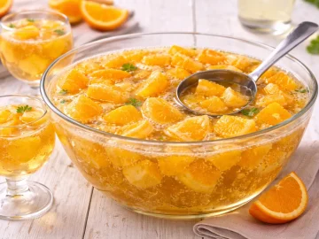 Glasgefäß mit Orangenbowle, Orangenstücken und Schöpfkelle auf weißem Holzhintergrund mit sichtbarer Maserung