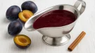Pflaumensoße in glänzender Edelstahlsauciere, serviert auf weißem Holzhintergrund mit frischen Pflaumen und Gewürzen.