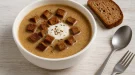 Thüringer Biersuppe in rustikaler Suppenschüssel, serviert mit Brotwürfeln und Sahne, auf hellem Holzhintergrund mit Besteck.