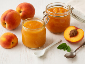 Pfirsichmus und Marmelade in Einmachgläsern, dekoriert mit frischen Pfirsichen und Löffel, auf hellem Holztisch