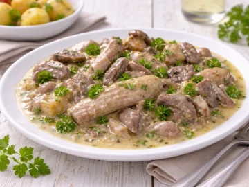 Weißer Teller mit Gänseklein in cremiger Sauce, Petersilie und Beilage auf weißem Holzhintergrund mit sichtbarer Maserung