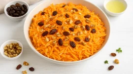 Frische Möhren-Apfel-Rohkost mit Nüssen und Rosinen in heller Schüssel auf weißem Holzuntergrund