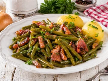 Teller mit grünen Bohnen und knusprigem Speck, garniert mit Petersilie auf hellem Holztisch