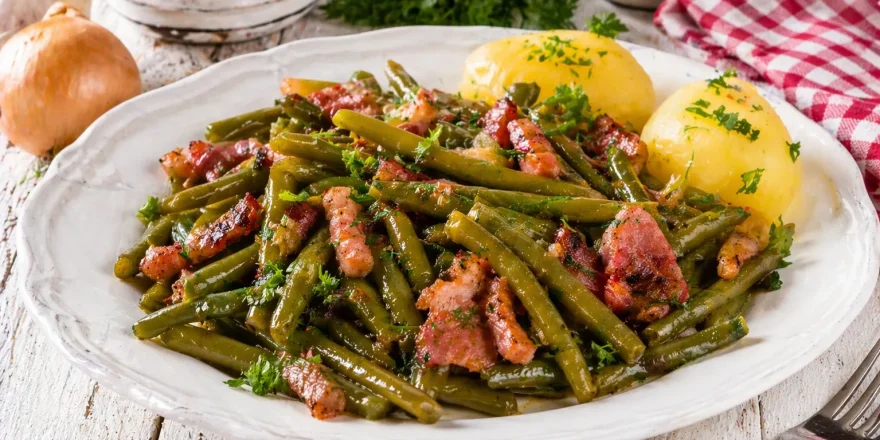 Teller mit grünen Bohnen und knusprigem Speck, garniert mit Petersilie auf hellem Holztisch