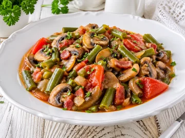 Buntes Gemüseragout mit grünen Bohnen, Pilzen und Tomaten in einer weißen Schüssel auf rustikalem Holztisch angerichtet