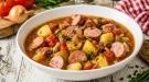 Gemüsetopf mit Bockwurst, Kartoffeln und Tomaten in weißer Schüssel auf hellem Holztisch angerichtet