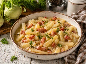 Kohlrabigemüse mit Speck in rustikaler Schüssel auf weißem Holztisch mit Kräutern und Küchentuch