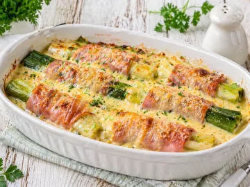 Überbackener Porree mit Schinken und cremiger Käsesoße in einer weißen Auflaufform auf hellem Holztisch angerichtet