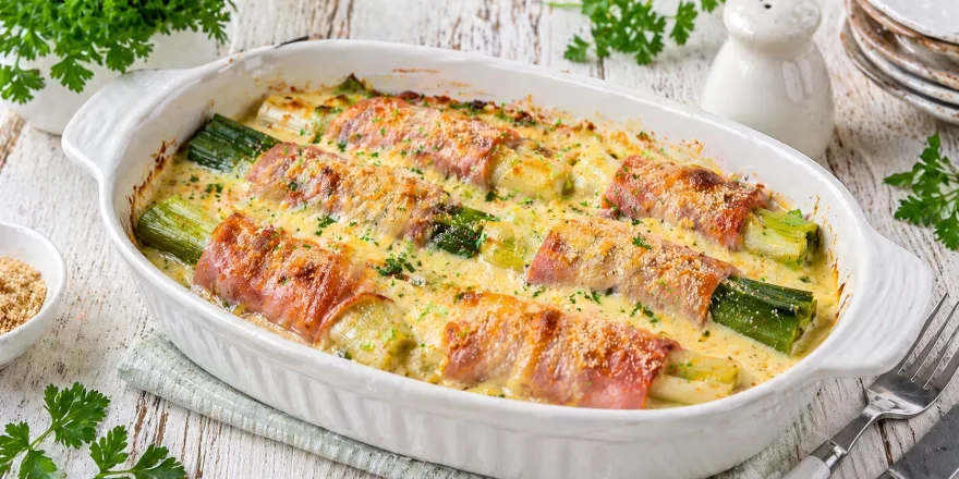 Überbackener Porree mit Schinken und cremiger Käsesoße in einer weißen Auflaufform auf hellem Holztisch angerichtet