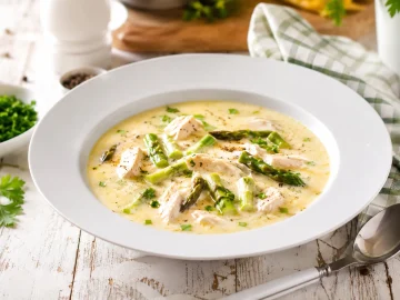 Cremige Hühnersuppe mit Spargelköpfen und zartem Hühnerfleisch in weißem tiefen Teller auf rustikalem Holztisch