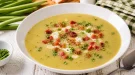 Cremige Bohnensuppe mit Speck und Petersilie in weißem tiefen Teller auf hellem Holztisch angerichtet