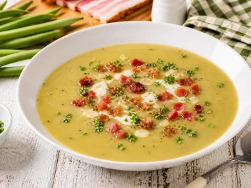 Cremige Bohnensuppe mit Speck und Petersilie in weißem tiefen Teller auf hellem Holztisch angerichtet