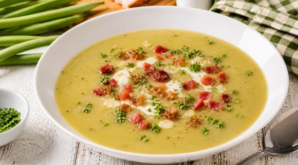 Cremige Bohnensuppe mit Speck und Petersilie in weißem tiefen Teller auf hellem Holztisch angerichtet