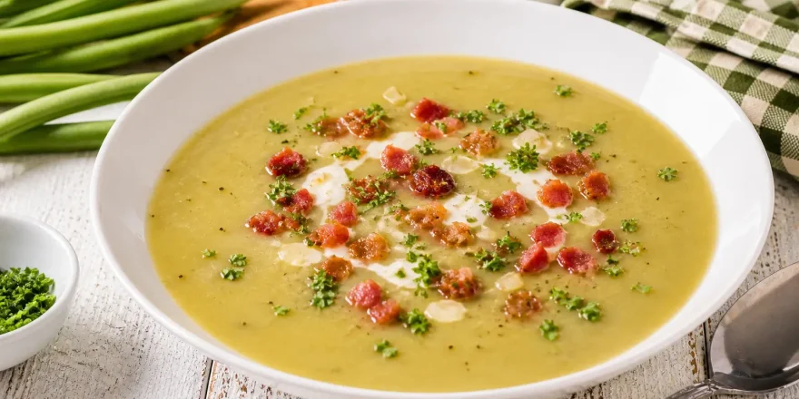 Cremige Bohnensuppe mit Speck und Petersilie in weißem tiefen Teller auf hellem Holztisch angerichtet