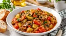 Weißer Teller mit Gemüseragout aus Pilzen, Gurken und Tomaten auf rustikalem weißem Holztisch