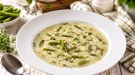 Cremige Grüne-Bohnen-Suppe in modernem weißen tiefen Teller auf rustikalem weißen Holztisch mit Kräutern und Besteck