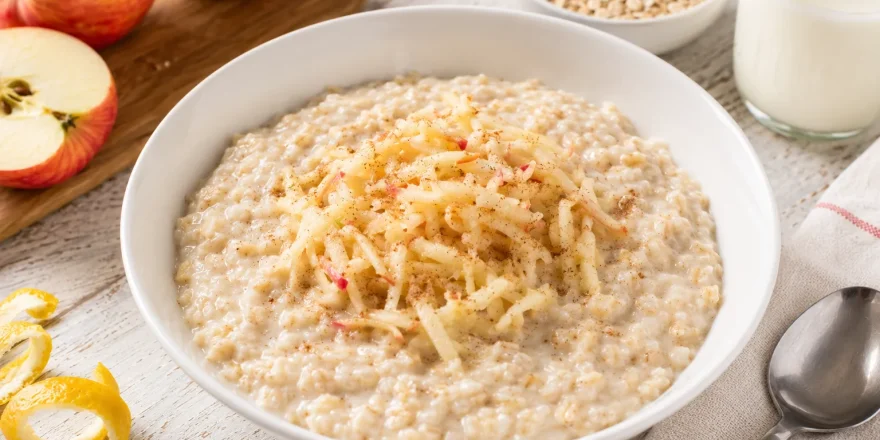 Cremiger Haferflockenbrei mit geriebenem Apfel in moderner weißer Schüssel auf rustikalem weißen Holztisch