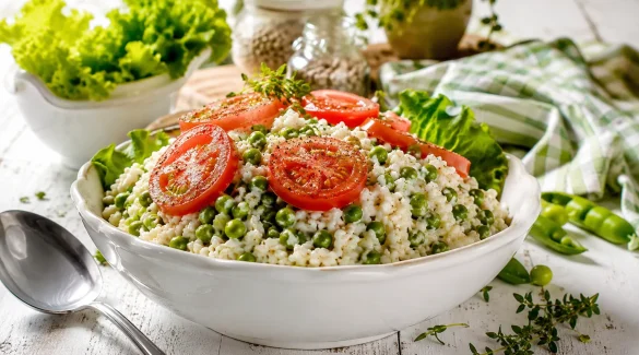 Weiße Schüssel mit cremigem Reissalat, grünen Erbsen und Tomatenscheiben auf rustikalem weißem Holztisch