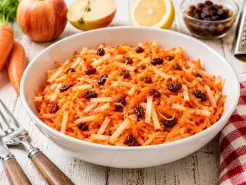 Frischer Möhrensalat aus geraspelten Möhren, Äpfeln und Rosinen in einer modernen weißen Schüssel.