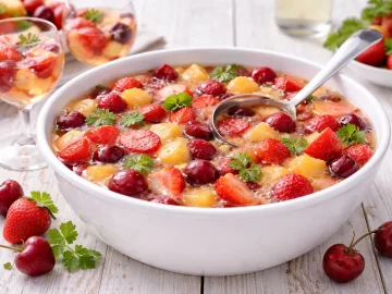 Weiße Bowlenschale mit Kinderbowle, Erdbeeren, Pfirsichen und Kirschen auf weißem Holzhintergrund mit sichtbarer Maserung und moderner Tischdeko