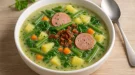 Cremige Kartoffelsuppe mit Gemüse, Spinat, Wurstscheiben, Speck und frischen Kräutern in einer weißen Suppenschüssel auf hellem Holzhintergrund.