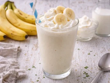 Bananen-Milchshake mit Vanilleeis und Milch im Glas mit Schlagcreme und Bananenscheiben auf weißem Holzhintergrund