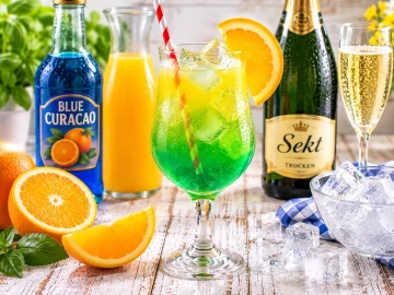 Grüne Wiese Cocktail aus Blue Curacao, Orangensaft und Sekt mit Eiswürfeln und Orangenscheibe in einem hohen Glas.