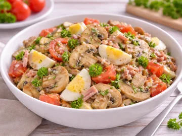 Pilzsalat mit Tomaten, Eiern, Schinken und Petersilie in einer weißen Salatschüssel auf hellem Holztisch
