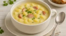 Kartoffelsuppe in einer weißen Suppenschüssel, serviert mit Petersilie, Würstchenscheiben und heller Tischdekoration