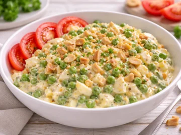 Cremiger Eiersalat mit grünen Erbsen, Sellerie, Tomaten und Nüssen in einer weißen Salatschüssel auf hellem Holztisch