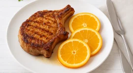 Gebratenes Kotelett mit Orangenscheiben auf einem weißen Teller, angerichtet auf hellem Holzhintergrund.